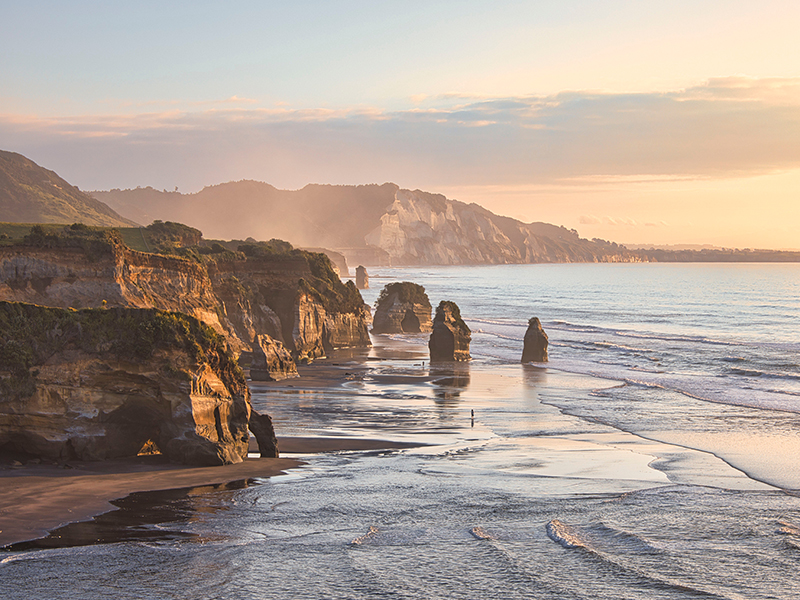 Three Sisters Rocks, New Zealand. Scenic view of a rugged coastline with tall sea stacks, gentle waves, and golden sunset light reflecting off the wet sand